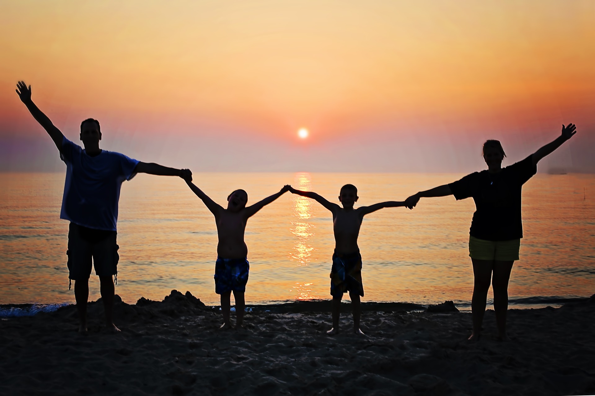 A family of four, a father, two kids, and a mother, hold hands and lift their arms in the air as they pose in front of a sunset on the water.