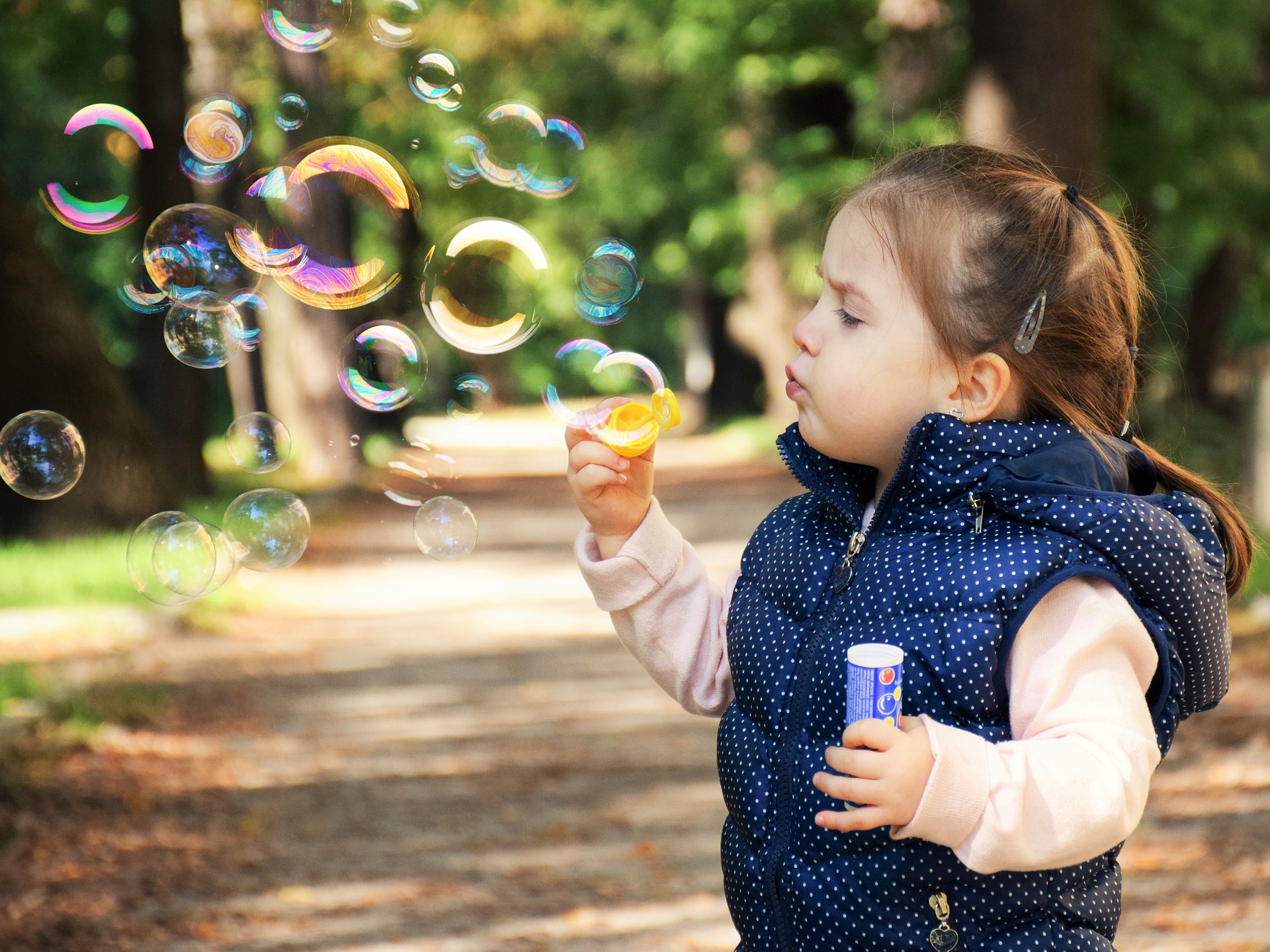 A child blows bubbles for fun on the sidewalk outside. She’s dressed in a pink sweatshirt and a blue with white polka dots gilet over it.